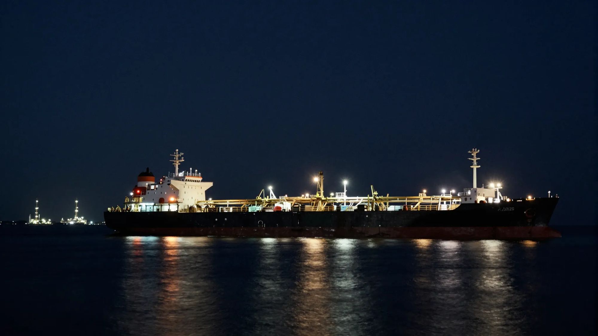 A crude oil tanker lit at anchor at night, refinery lights on the horizon.